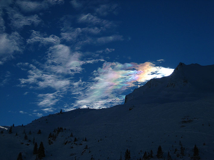 A cloud reflecting rainbow colors in the blue sky behind a snowy mountain.