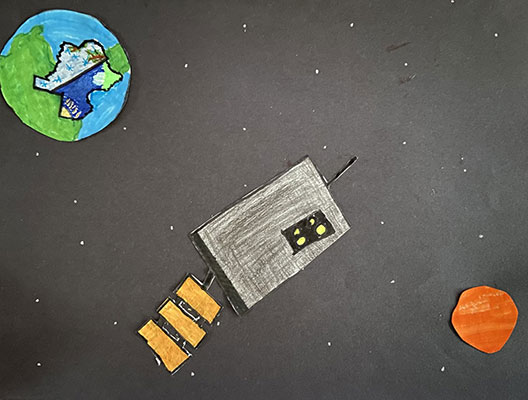 Paper cutouts of Earth, GOES-T and Mars on a black piece of paper.