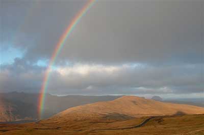 Photo of rainbow arc over brown hills, with road.