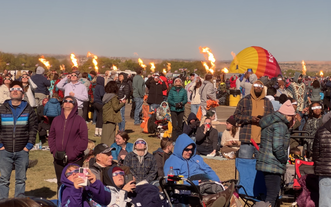 Image of a group of people looking at an eclipse with hot air ballons around them.