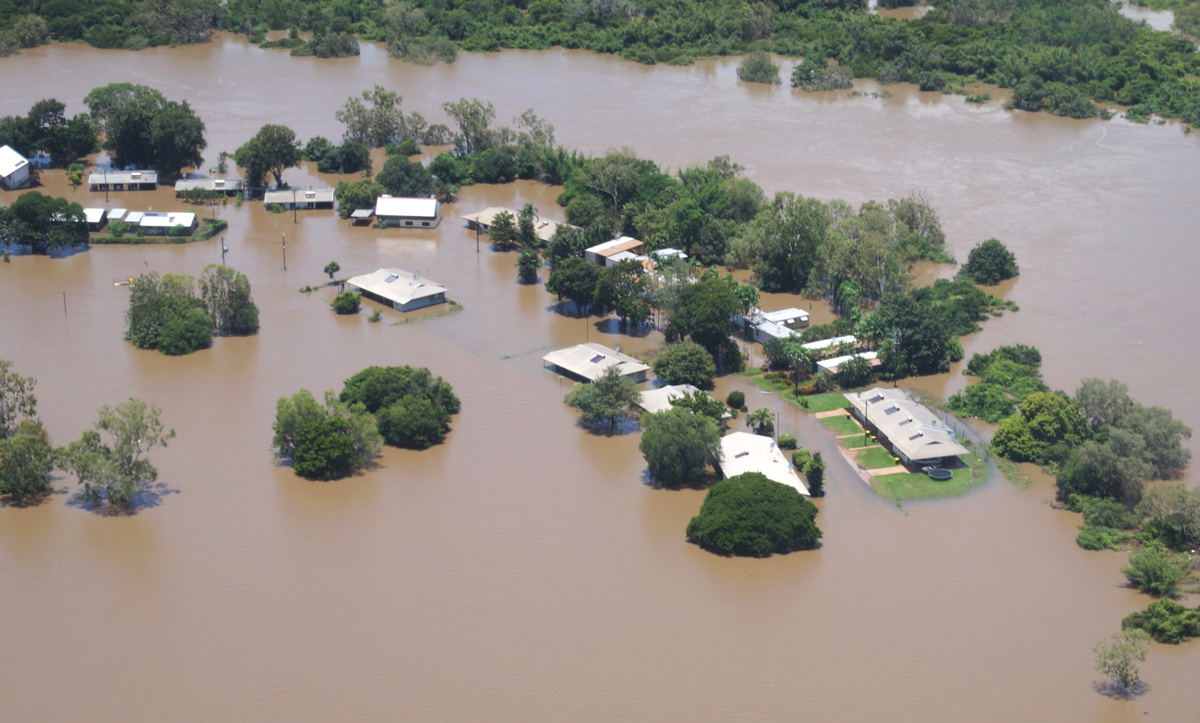Aerial view of a flooded residential area with homes partially submerged, surrounded by muddy floodwaters and scattered greenery.