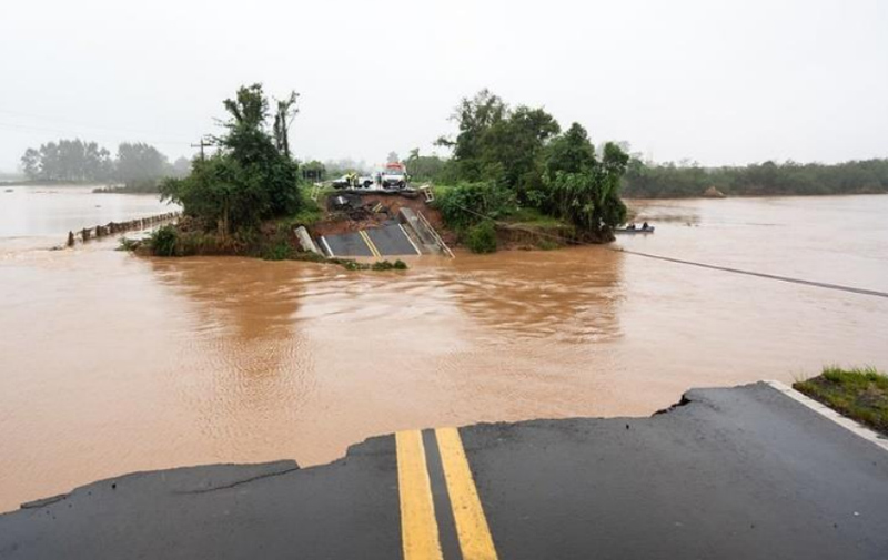 A collapsed road and submerged bridge in Rio Grande do Sul, Brazil, after extreme flooding, with muddy waters flowing under a damaged section and trees in the background.