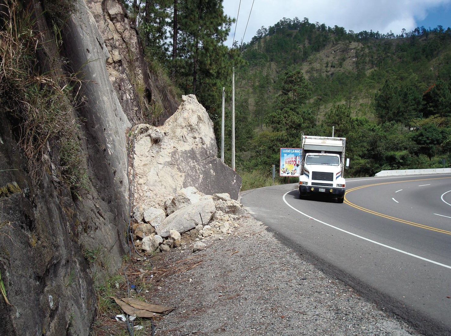 A landslide partially blocking a roadside with a large rock mass, located near a curved mountain road as a truck passes by, under a cloudy sky. 