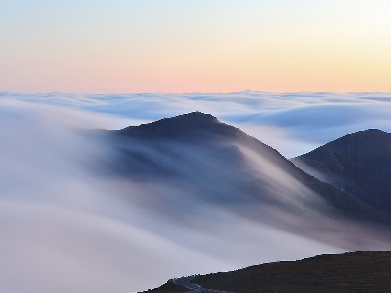 Light gray clouds flowing over dark mountain peaks with a soft pink sky above