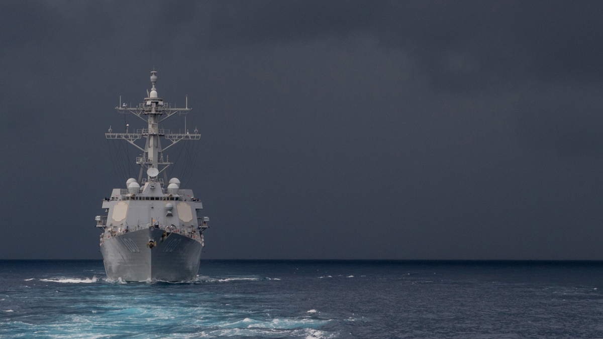Navy ship sails through dark ocean waters under a stormy sky, leaving a foamy wake behind. The vessel is heading forward, facing turbulent weather.