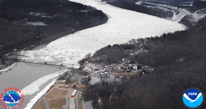 Aerial view of an ice-jammed river flooding a small town, with NOAA and NWS logos in the corners.