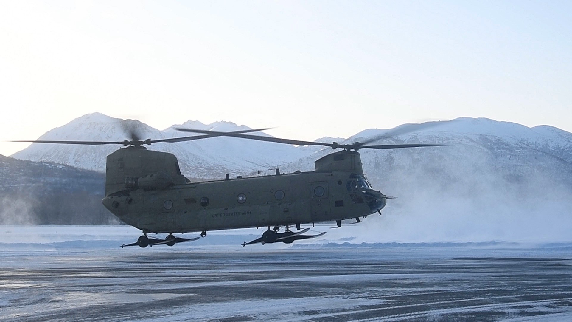 U.S. Army CH-47 Chinook helicopter hovers over a snowy runway with mountains in the background and snow blowing from rotor wash.