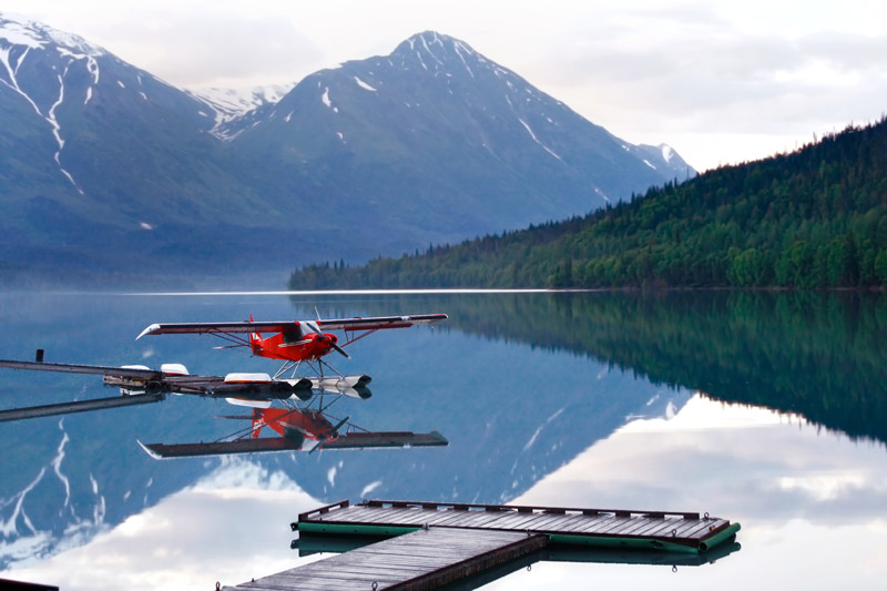 A red floatplane is moored on a calm, glassy lake, reflecting the plane, a wooded shoreline, and snow-dusted mountains.
