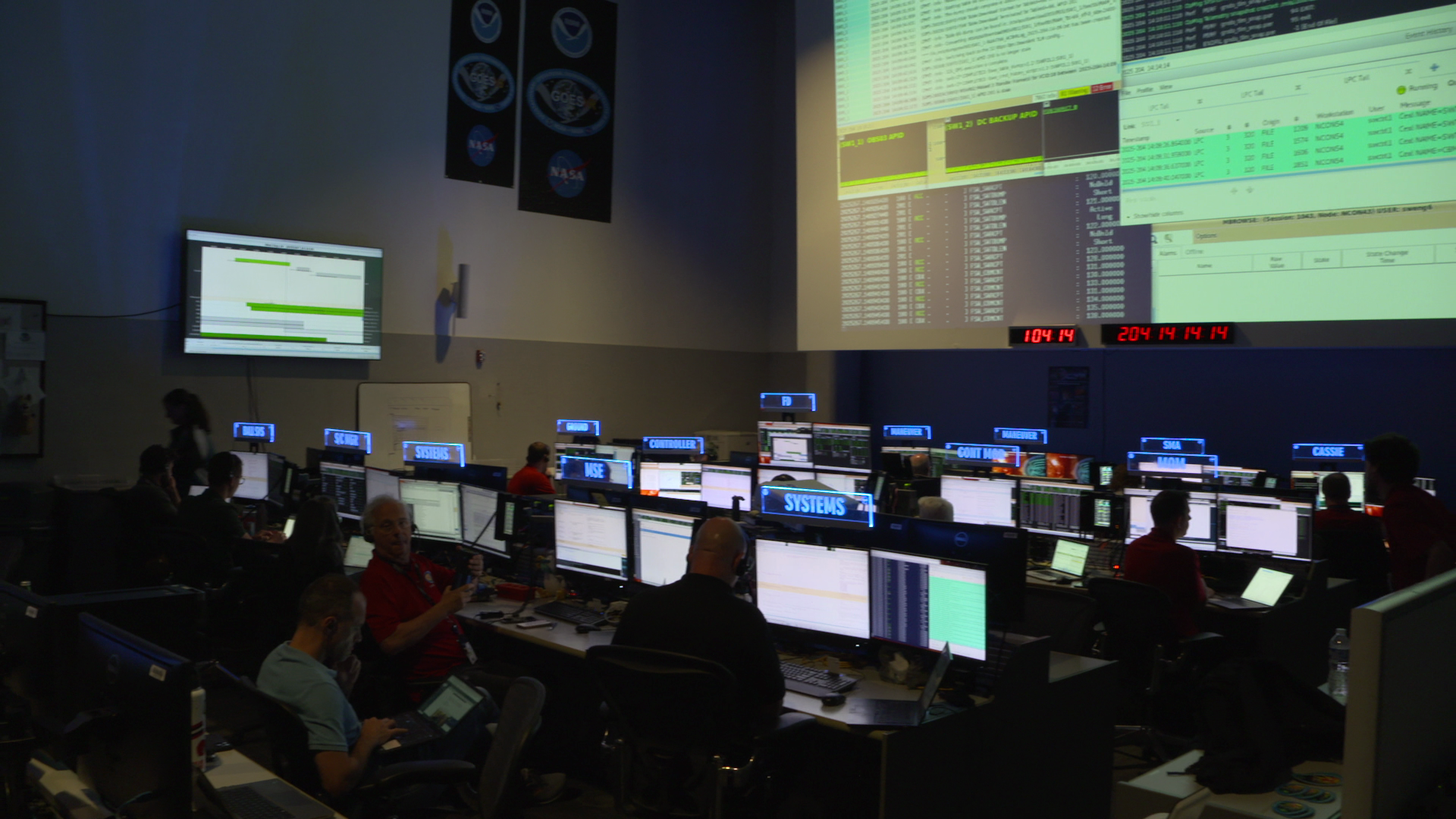 A room with large screens and rows of computers and cubicle.