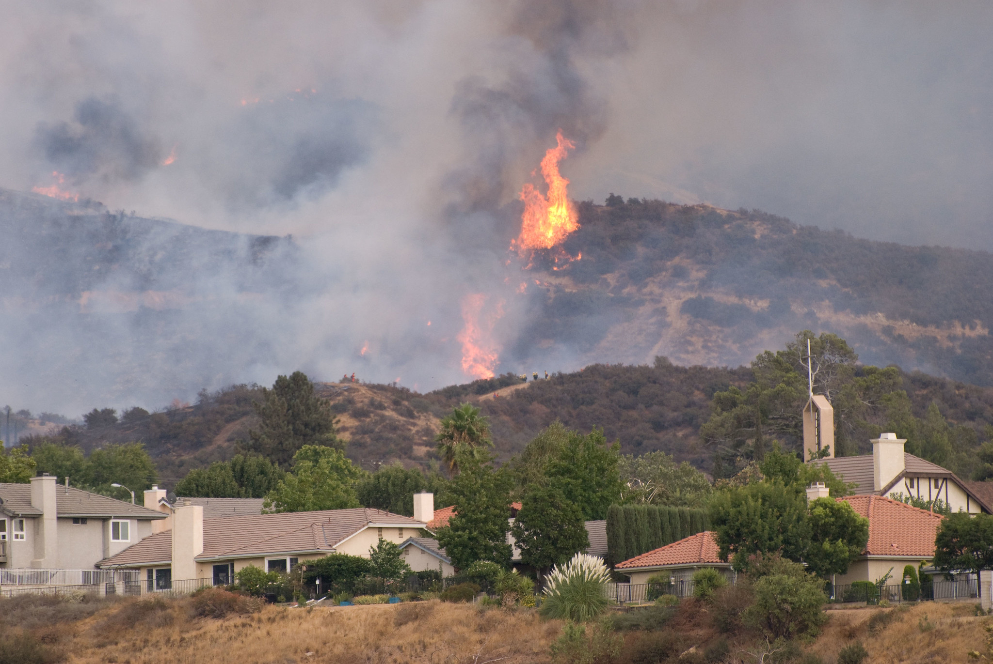 A wildfire burns intensely on a dry, brush-covered hill behind a suburban neighborhood with visible flames and thick smoke threatening the homes below.