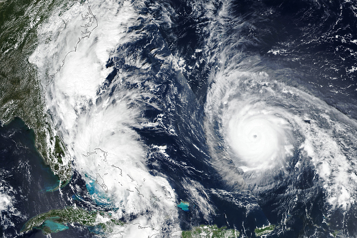 Satellite image of two large white swirling cloud masses over the ocean. The right storm has a clear eye. The left storm is closer to a green coastline.
