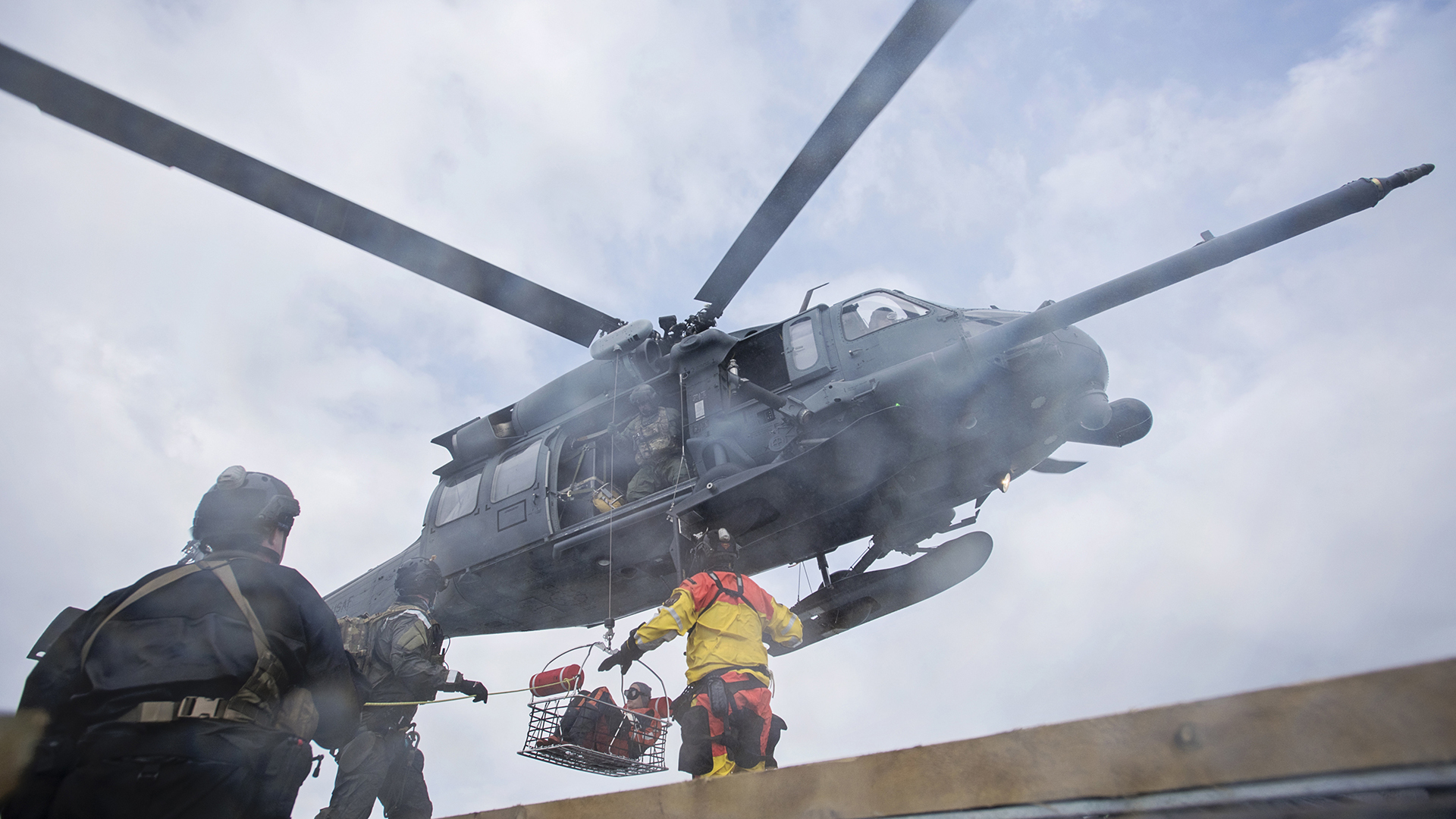 Low-angle shot of a military helicopter hovering as personnel hoist a person in a rescue basket from a deck during a maritime extraction.