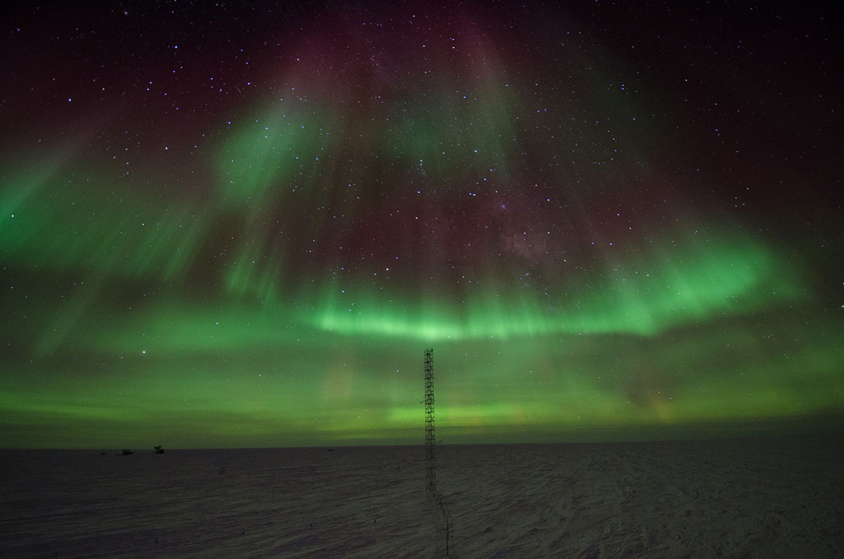 Green horizontal streaks of light illuminate a dark sky with a thin tower in the foreground.