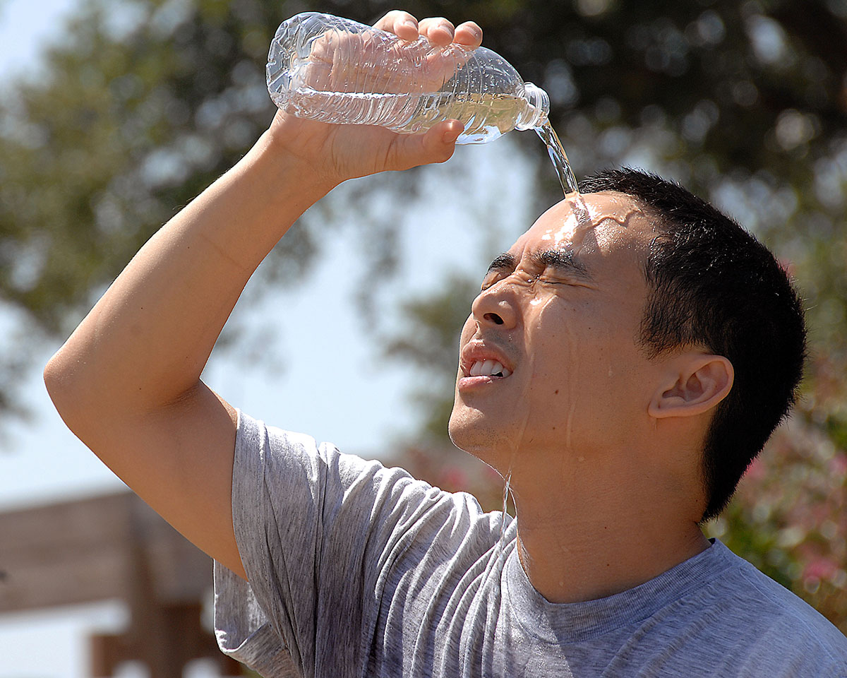 A man in a light-gray t-shirt is outside, holding an open plastic water bottle over his head, pours the remaining water inside the bottle over his forehead to cool off in the humid heat. The water runs down his face and onto his shirt.